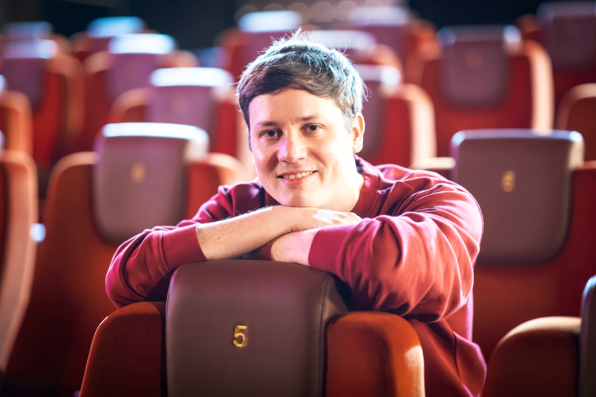 David Reiter sitzt in einem leeren Kinosaal. Er lehnt mit verschränkten Armen auf der Lehne des Vordersitzes und blickt in Richtung Leinwand und Kamera.