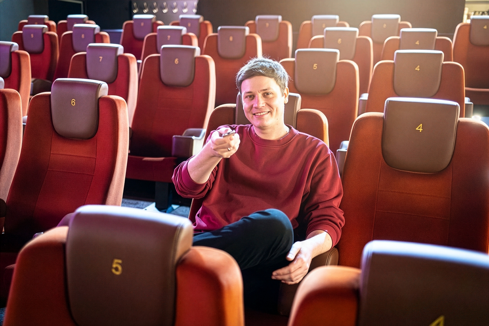 David Reiter sitzt mittig in einem leeren aber gut beleuchteten Kinosaal und zeigt mit einer Streaming-Fernbedienung lächelnd in Richtung Leinwand und Kamera.